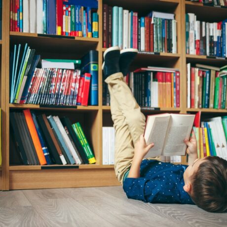 Băiat stă pe podea cu picioarele pe bibliotecă și citește