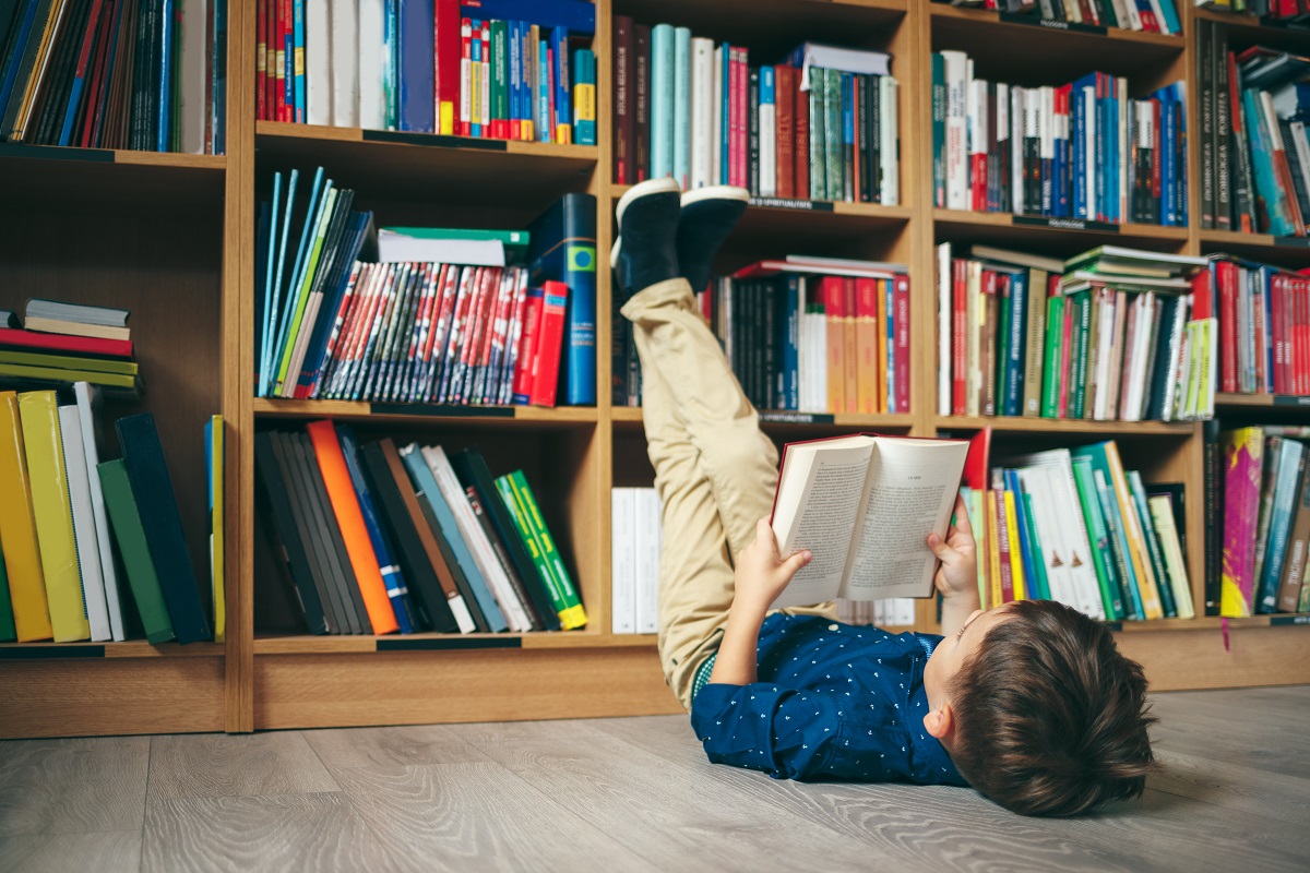 Băiat stă pe podea cu picioarele pe bibliotecă și citește