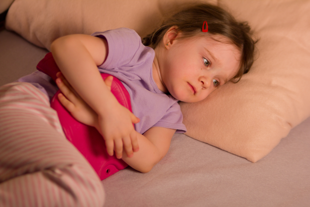 Fetiță mică stă în pat tristă, cu o pernă pe burtă
