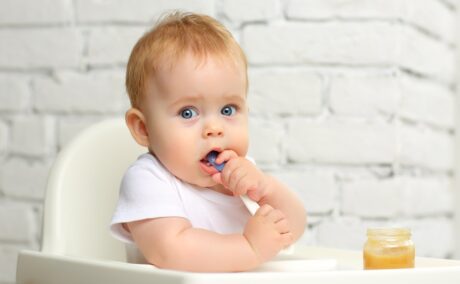 Un bebeluș care stă într-un scaun special și mănâncă dintr-un borcănaș în care se află o pastă de cartofi dulci