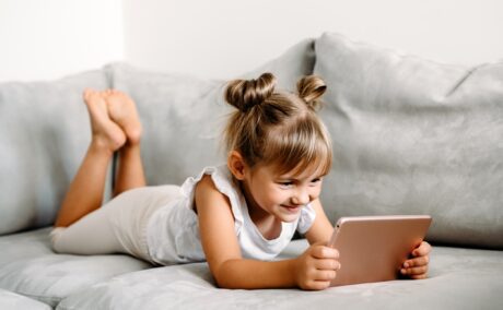 O fetiță cu codițe care stă pe o canapea și se joacă pe tabletă pentru a ilustra cât de nociv este timpul petrecut de un copil în fața ecranelor