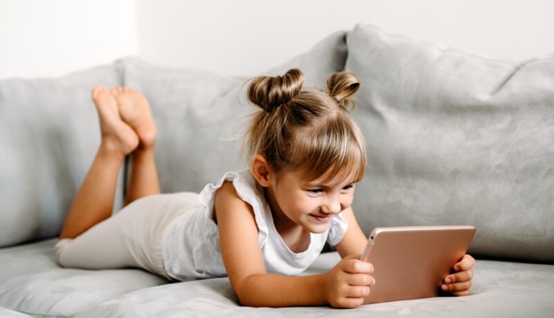 O fetiță cu codițe care stă pe o canapea și se joacă pe tabletă pentru a ilustra cât de nociv este timpul petrecut de un copil în fața ecranelor