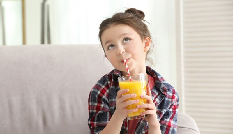 O fetiță frumoasă care bea suc de portocale dintr-un pahar folosind un pai colorat