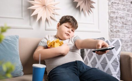 Băiat cu greutate peste medie stă pe canapea cu o telecomandă în mână