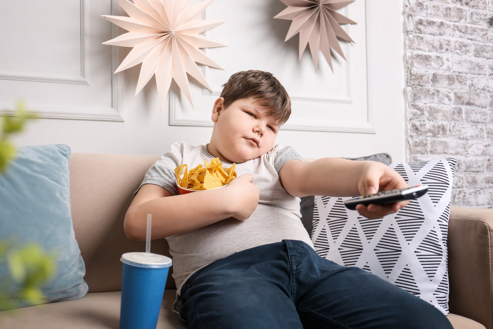 Băiat cu greutate peste medie stă pe canapea cu o telecomandă în mână