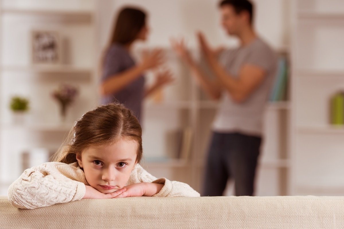 Fetița stă tristă pe canapea în timp ce părinții se ceartă în spatele ei