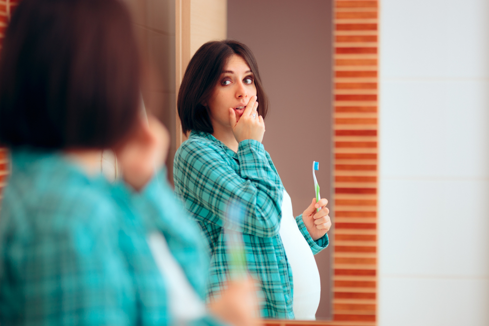 Femeie însărcinată ține o periuță de dinți în mână și se uită seriată în oglindă