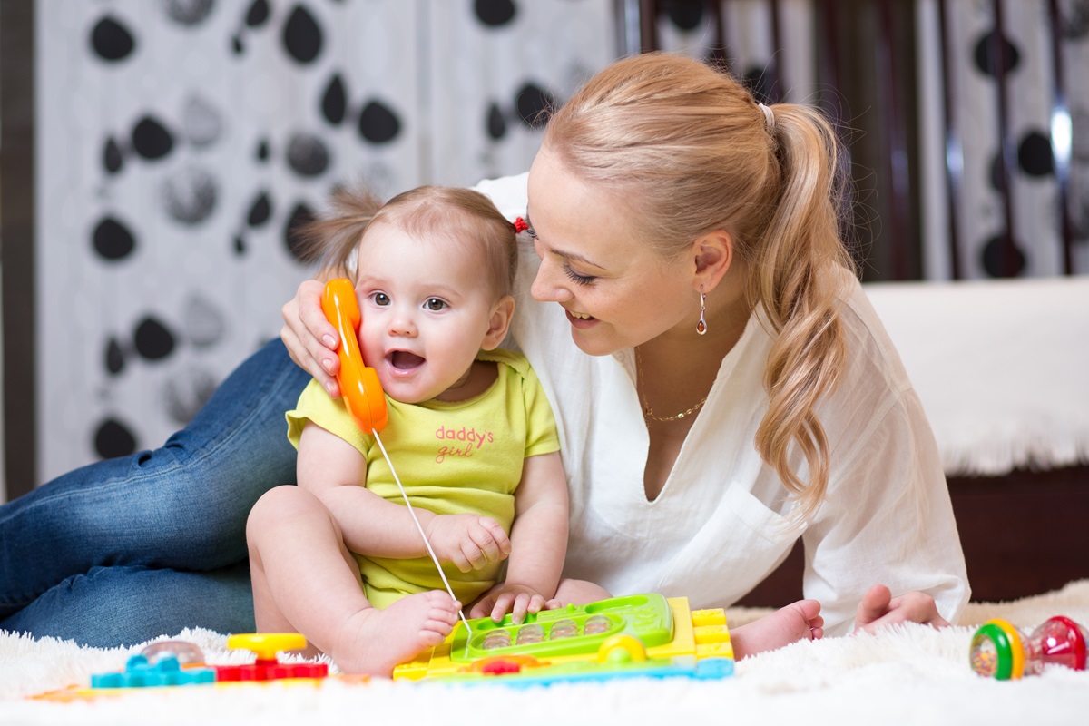 O mămică care stă pe un covor alături de fiica ei în timp ce îi ține la ureche un telefon de jucărie pentru a ilustra motivul pentru care unii copii învață să vorbească mai repede decât alții