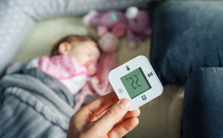 O mămică care ține un termometru deasupra pătuțului copilului ei pentru a arăta cum îți dai seama că bebelușului tău îi este prea cald