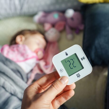 O mămică care ține un termometru deasupra pătuțului copilului ei pentru a arăta cum îți dai seama că bebelușului tău îi este prea cald