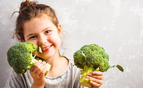 O fetiță frumoasă care ține în mână două buchețele cu broccoli, bogate în vitamine esențiale