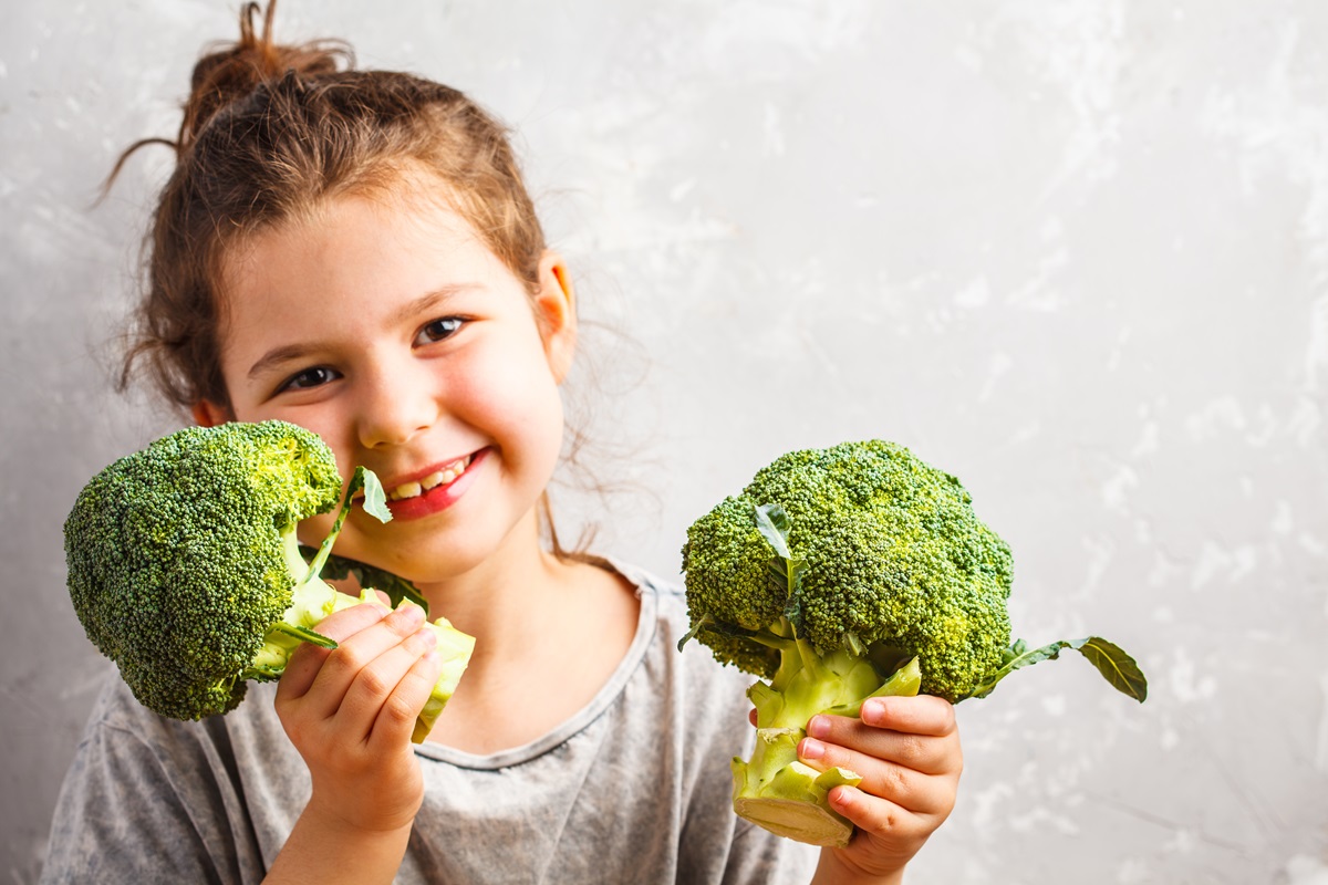 O fetiță frumoasă care ține în mână două buchețele cu broccoli, bogate în vitamine esențiale
