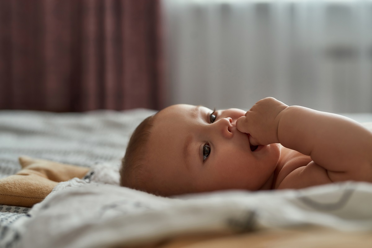 Bebelușul stă pe pat întins pe spate cu degetul în gură