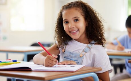 Fetiță care stă în bancă la școală și zâmbește cu creionul în mână
