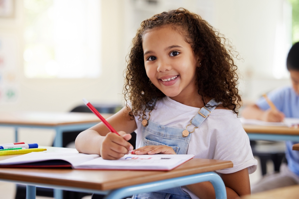 Fetiță care stă în bancă la școală și zâmbește cu creionul în mână