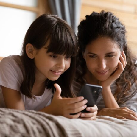 O mamă și o fiică în timp ce stau pe o saltea și se uită împreună la un telefon pentru a ilustra cum să-ți ajuți copilul născut în era digitală