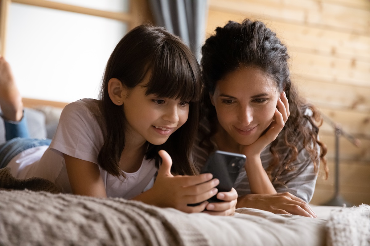 O mamă și o fiică în timp ce stau pe o saltea și se uită împreună la un telefon pentru a ilustra cum să-ți ajuți copilul născut în era digitală