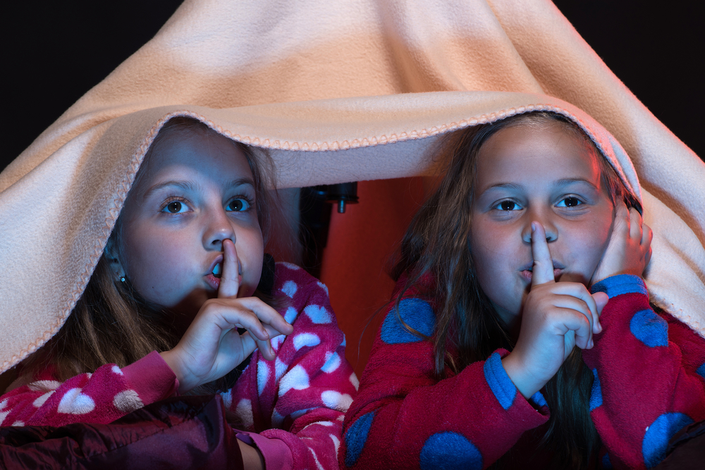 Două fetițe care stau sub o pătură, cu lumina stinsă