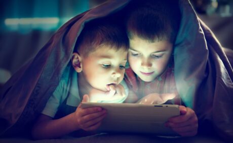 Doi copii care se joacă împreună pe o tabletă în timp ce se ascund de părinți sub o pătură