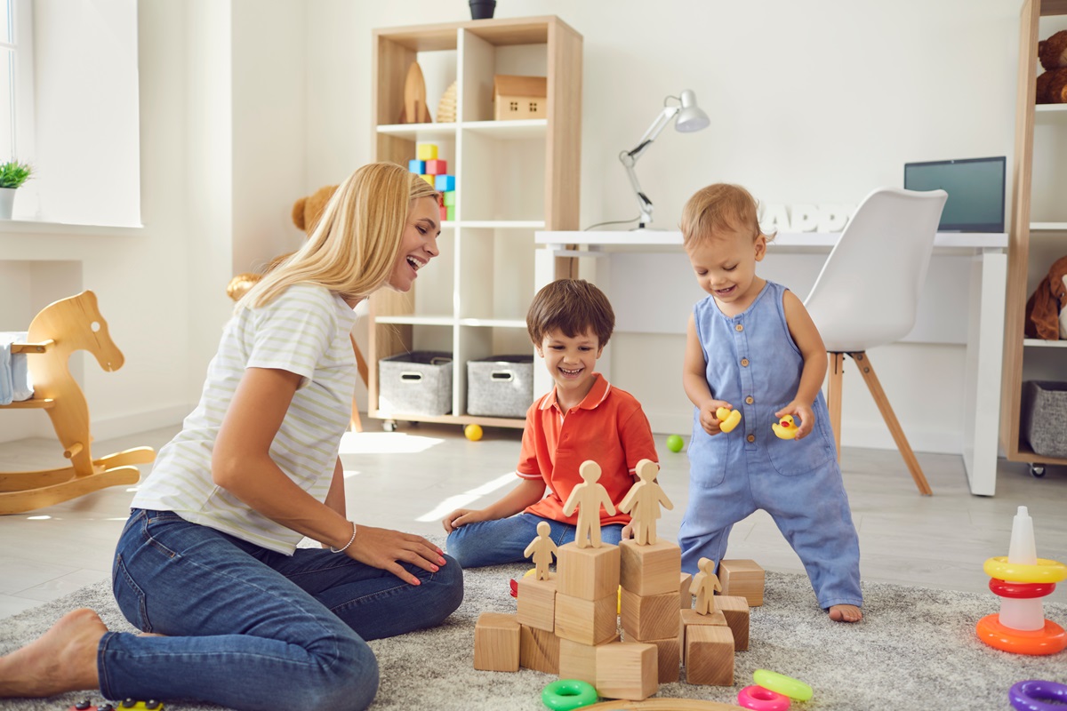 O mămică ce se joacă cu doi copii într-o cameră plină cu jucării de lemn pentru a ilustra modul în care jocurile pot să îmbunătățească dispoziția unui copil