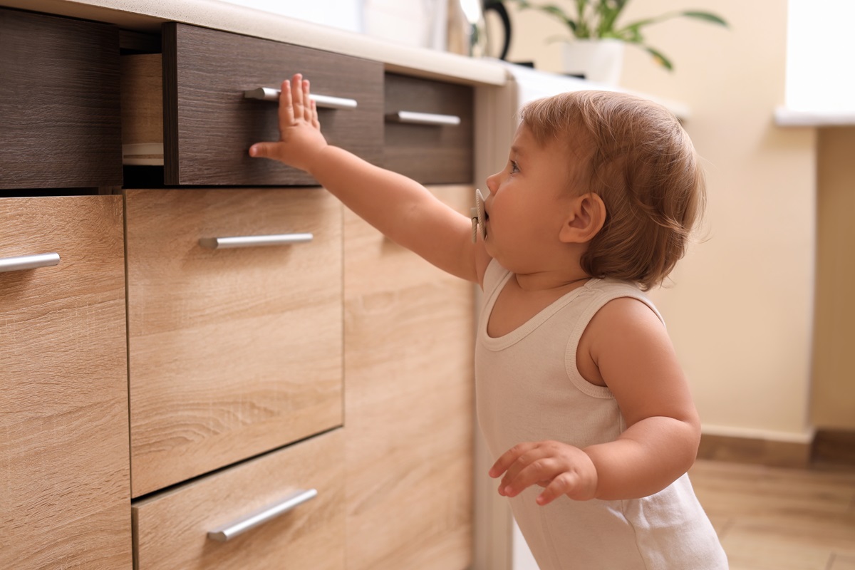 Un copil curios care ține o suzetă în gură și împinge cu putere ăn sertarul de la un corp de bucătărie