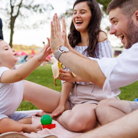 Tatăl se joacă cu bebelușul la picnic, în timp ce mama îi dă o gustare