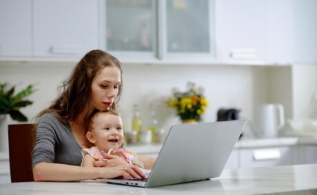 O mămică care ăși ține copilul în brațe în timp ce citește de pe laptop câteva adevăruri inconfortabile despre parenting