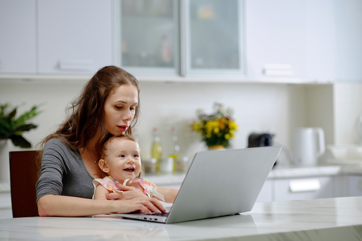 O mămică care ăși ține copilul în brațe în timp ce citește de pe laptop câteva adevăruri inconfortabile despre parenting