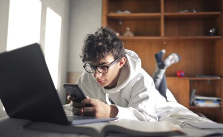 Adolescent stă întins în pat cu telefonul în mână și laptopul lângă el