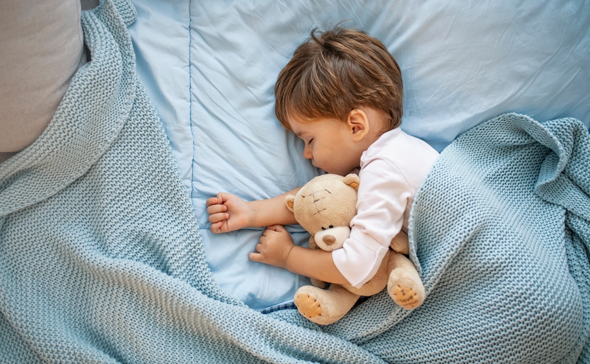 Un băiețel care doarme învelit cu o păturică albastră în timp ce își ține în brațe ursulețul