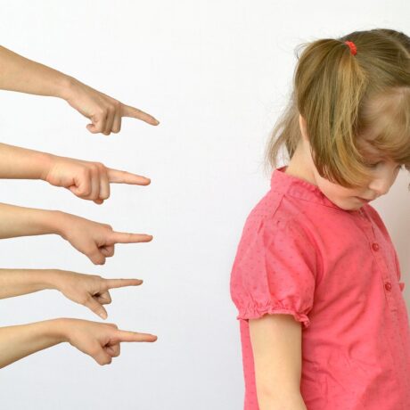 Fetiță stă supărată în timp ce mai multe mâini o arată cu degetele