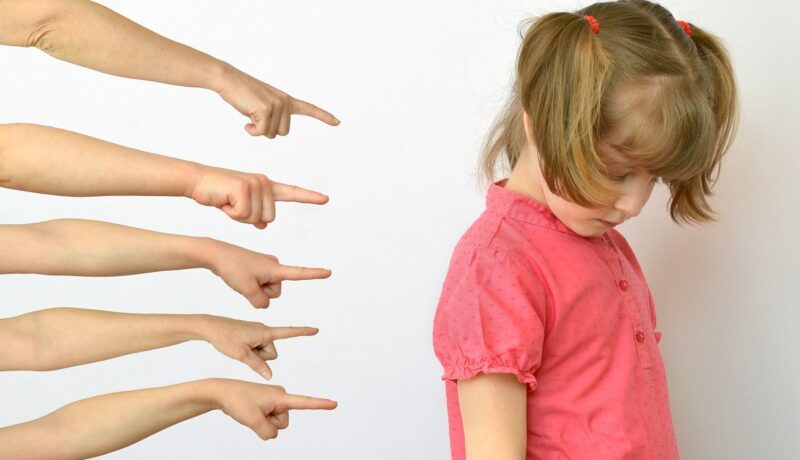 Fetiță stă supărată în timp ce mai multe mâini o arată cu degetele