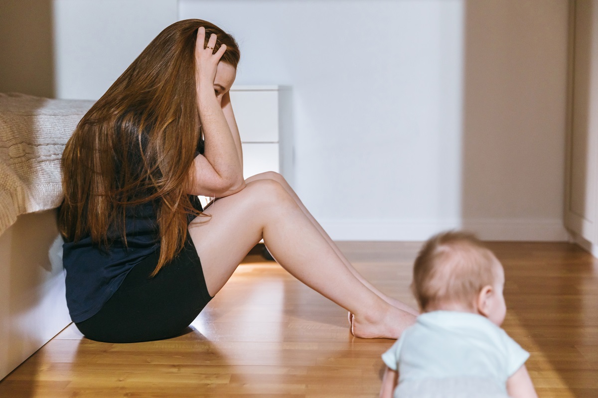 Mama stă pe podea cu mâinile la cap, în timp ce copilul se joacă pe lângă ea