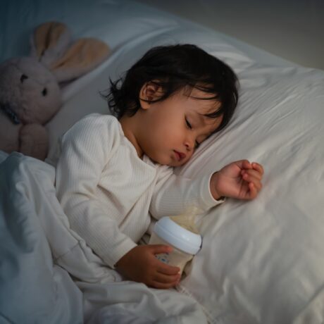 Un toddler care doarme în pătuț în timp ce ține biberonul în mână