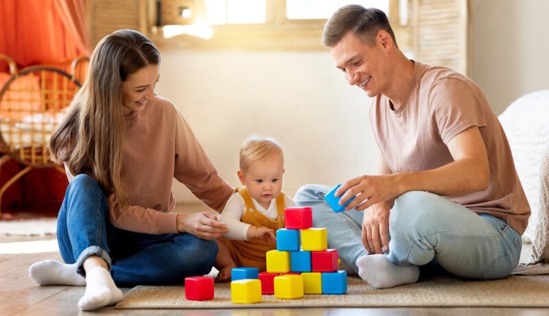 o mamă și un tătic care se joacă cu bebelușul pe un covor și construiesc un turn din cuburi colorate