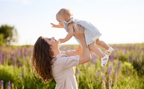 Mama se află cu fetița pe un câmp de flori și o ridică pe micuță în sus