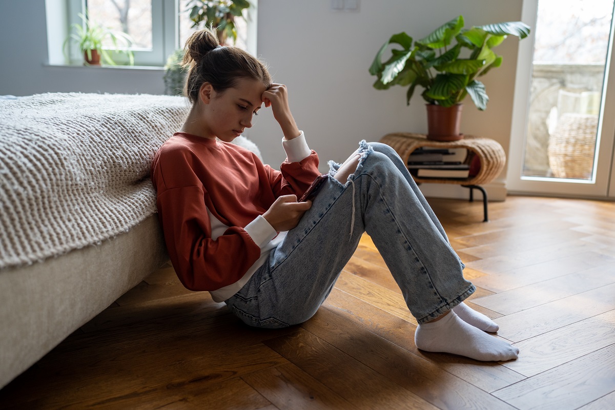 Adolescenta stă supărată pe podea, cu telefonul în mână