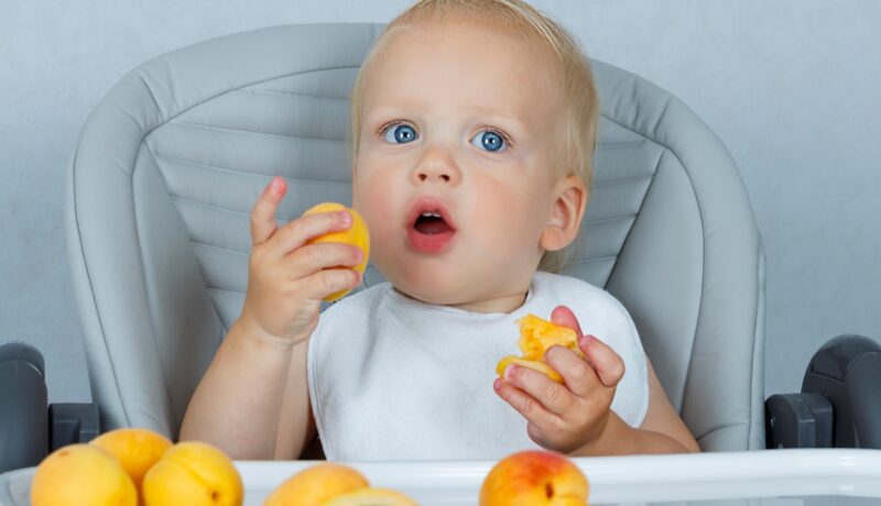 Un bebeluș care stă la măsuță și consumă bucăți mici de caise