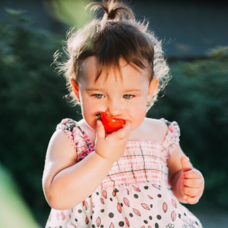 O fetiță care stă pe iarbă și mănâncă cu poftă o roșie