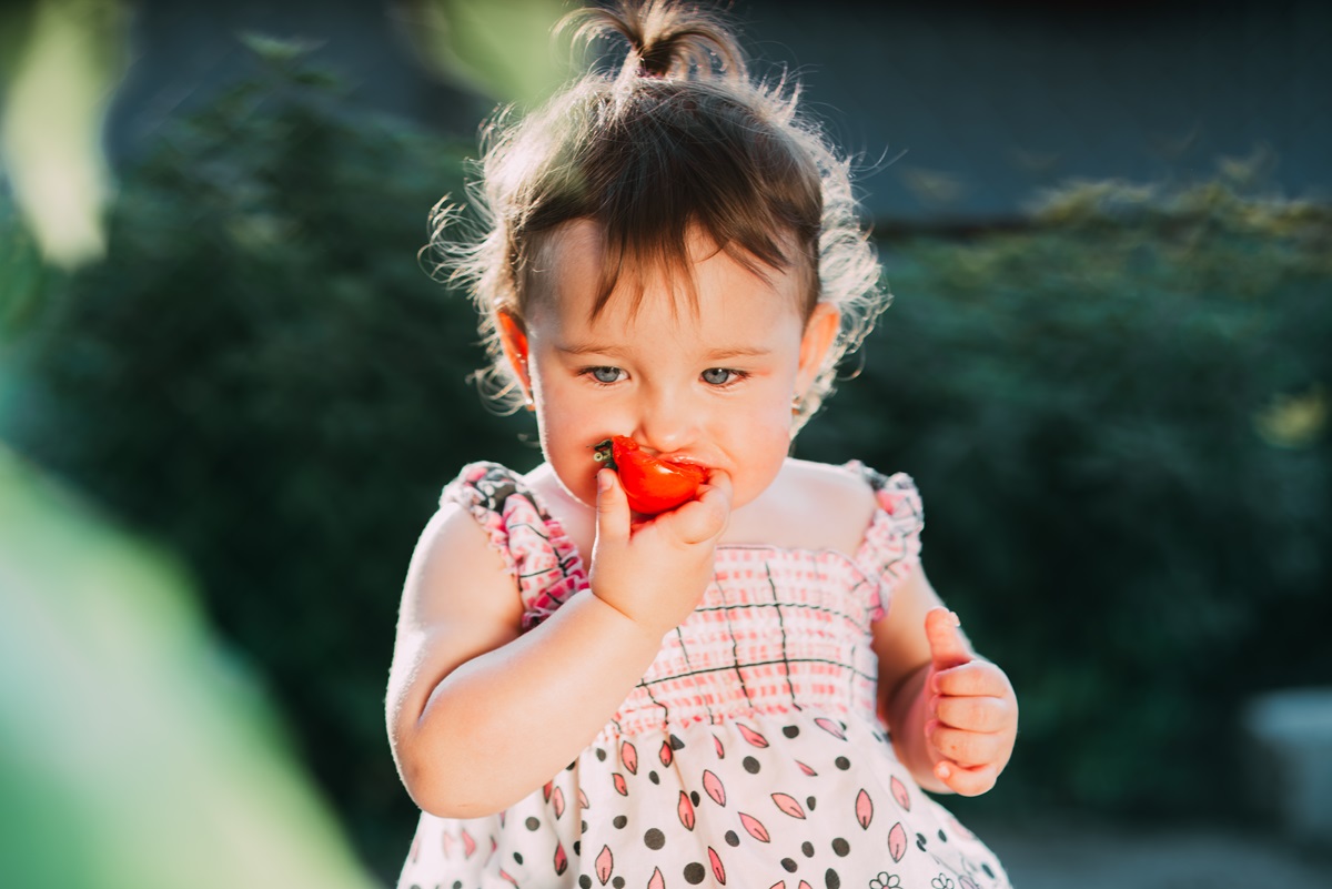 O fetiță care stă pe iarbă și mănâncă cu poftă o roșie
