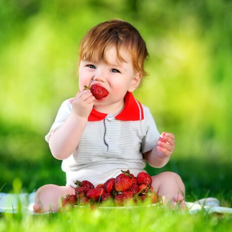 Un bebeluș care stă pe iarbă și mănâncă o căpșună mare și suculentă