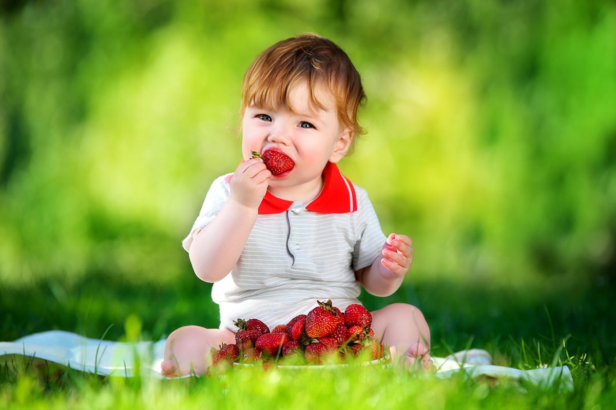 Un bebeluș care stă pe iarbă și mănâncă o căpșună mare și suculentă