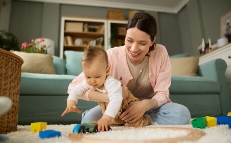 Mama stă cu bebelușul în brațe pe covor și se joacă