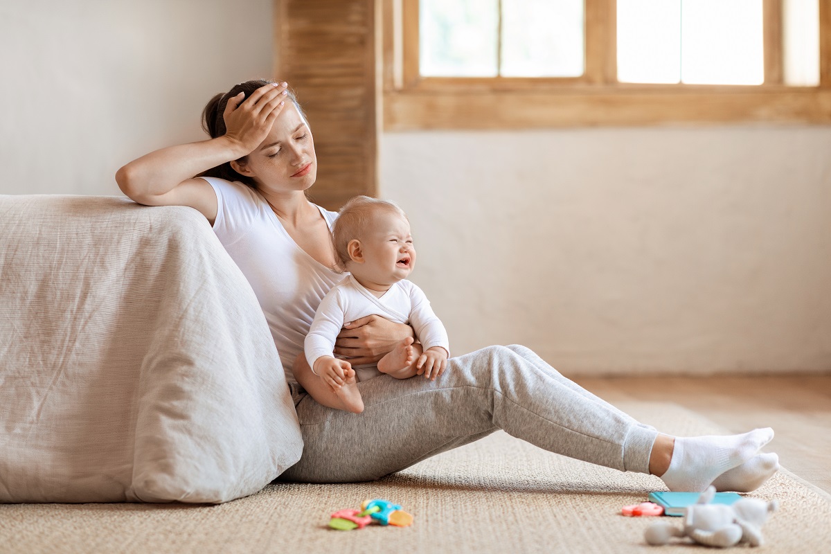 Mama stă cu bebelușul care plânge pe podea și este vizibil tristă și obosită