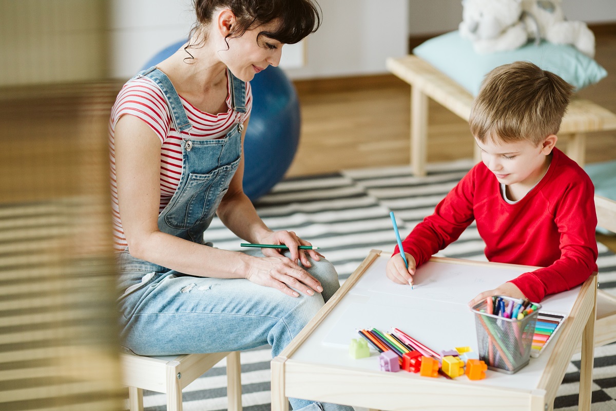 Mama stă pe scaun și supraveghează copilul care desenează