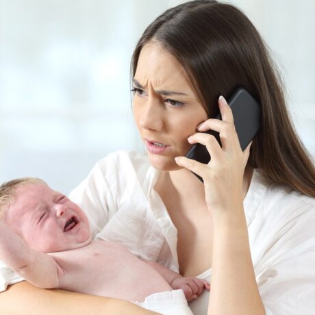 Mama vorbește îngrijorată al telefon în timp ce-și ține bebelușul care plânge în brațe