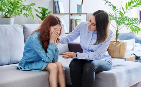 o mamă care stă pe canapea și își mângâie fiica care își ține fața în palme și plânge