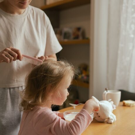 Mama îi aranjează fetiței părul în timp ce ia micul dejun