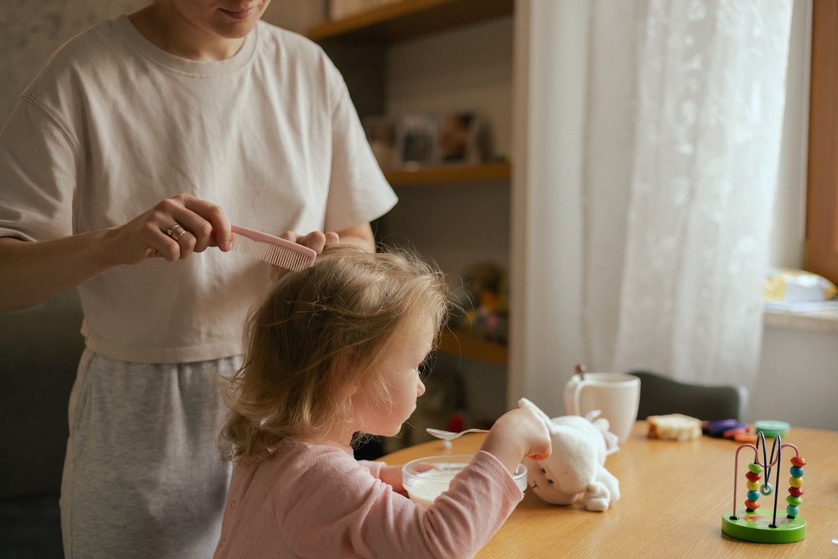 Mama îi aranjează fetiței părul în timp ce ia micul dejun