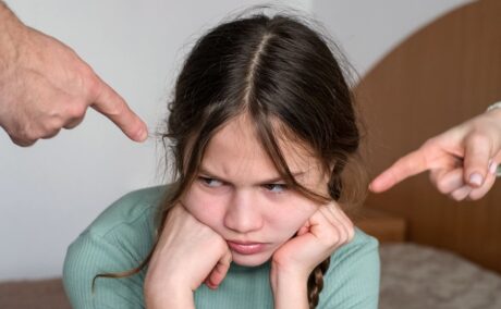 Fetiță stă supărată în timp ce este atenționată de mâinile părinților